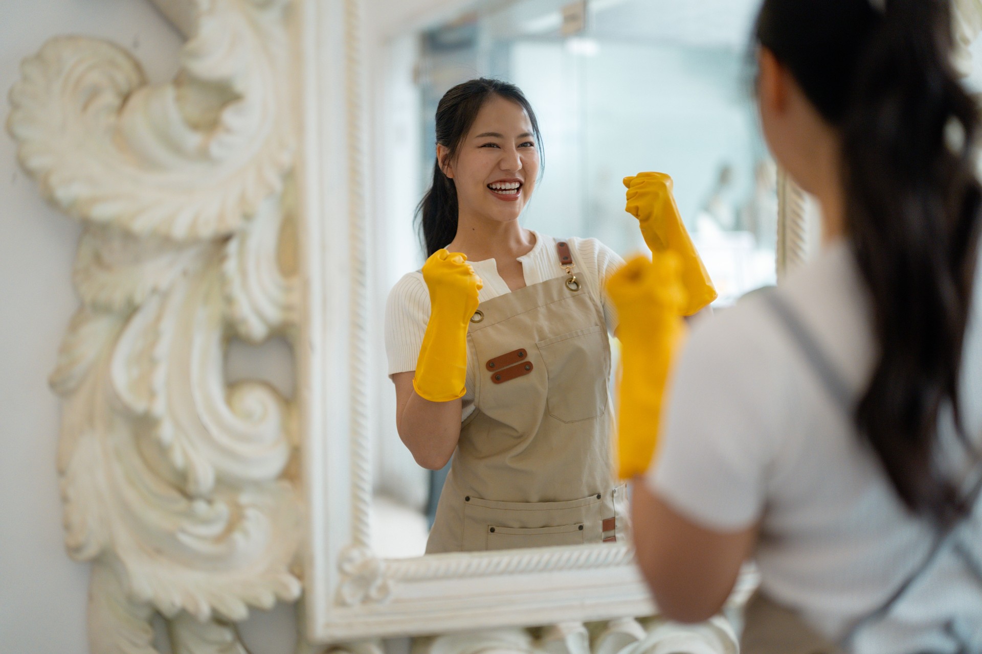 Happy housekeeper wearing yellow protective gloves and apron looking at the mirror and making winning gesture